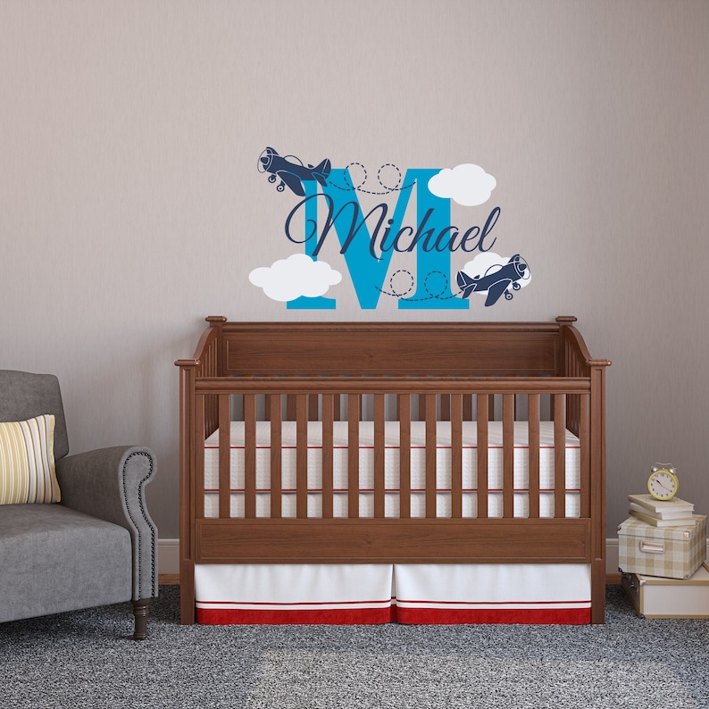 May include: A blue letter M with the name Michael written in a cursive font. Two blue airplanes are flying around the letter. White clouds are behind the letter. The decal is on a wall above a wooden crib with a white and red skirt.