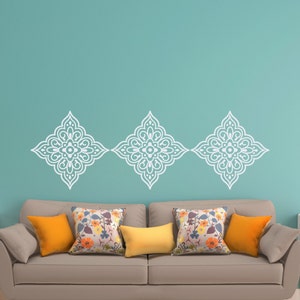 May include: Three white decorative wall decals in a diamond shape with a floral pattern, positioned above a light brown sofa with yellow and floral patterned throw cushions.