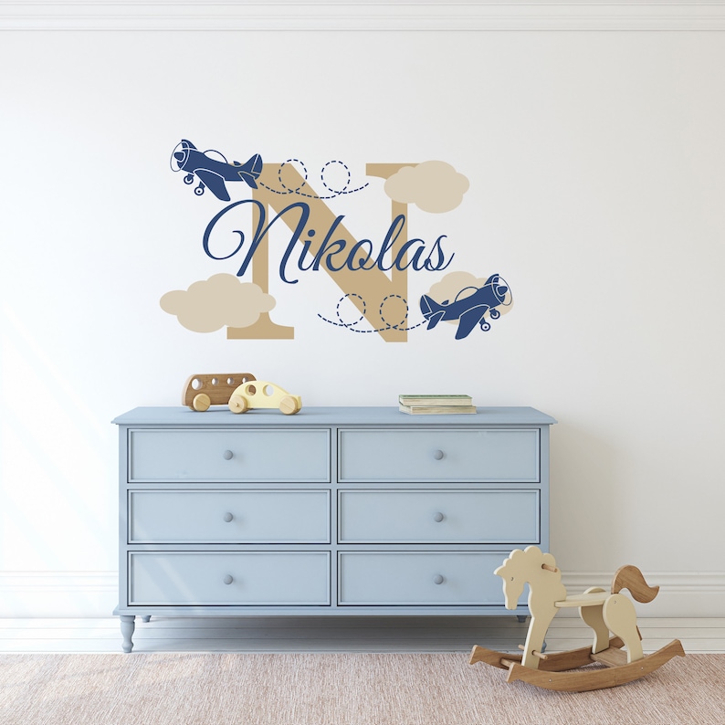 May include: A blue and tan nursery wall decal with the letter N and the name Nikolas. Two blue airplanes are flying above the letter with tan clouds.