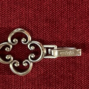 May include: A silver-toned bracelet link featuring a four-lobed, openwork clover design with curled edges. The piece includes a rectangular, hinged clasp mechanism attached to the side, showing a metallic finish and a smooth, polished surface.