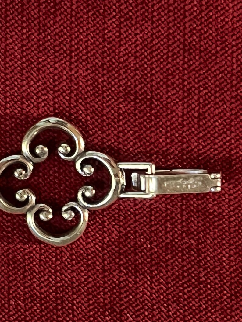 May include: A silver-toned bracelet link featuring a four-lobed, openwork clover design with curled edges. The piece includes a rectangular, hinged clasp mechanism attached to the side, showing a metallic finish and a smooth, polished surface.