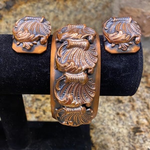 May include: A vintage copper-toned jewelry set featuring a wide cuff bracelet and matching square clip-on earrings. The set showcases an ornate, raised floral and scroll design with a dark patina in the recessed areas. The bracelet is displayed on a black velvet stand.