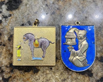 May include: Two vintage pendants. One is gold-toned, square, and features a horse with a rider. The other is blue and gold, with a figure holding a lantern. Both have small loops for hanging.