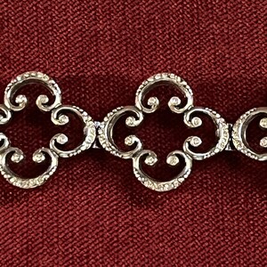 May include: A silver-toned metal bracelet featuring a repeating pattern of interconnected, openwork quatrefoil links. Each link displays a textured, granular surface finish along the curved edges, creating a consistent, linked chain design.
