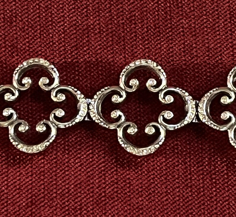 May include: A silver-toned metal bracelet featuring a repeating pattern of interconnected, openwork quatrefoil links. Each link displays a textured, granular surface finish along the curved edges, creating a consistent, linked chain design.
