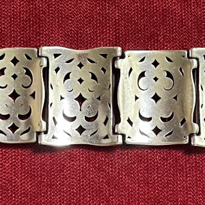 May include: A silver-toned metal bracelet featuring a series of rectangular links with an intricate, symmetrical cut-out pattern. The bracelet includes a rectangular buckle-style clasp on the left side, with each link connected by hinges.
