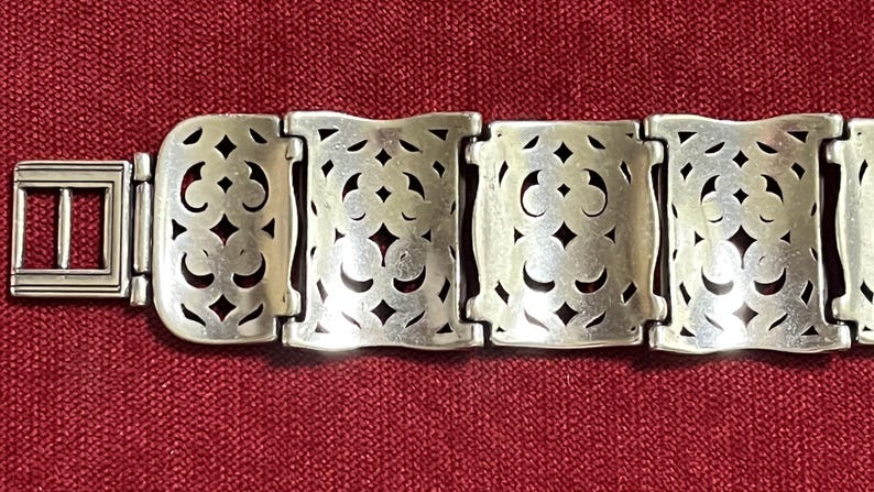 May include: A silver-toned metal bracelet featuring a series of rectangular links with an intricate, symmetrical cut-out pattern. The bracelet includes a rectangular buckle-style clasp on the left side, with each link connected by hinges.