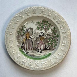 May include: A white ceramic plate with a green and brown illustration of two women in a garden. The plate is decorated with the alphabet around the edge.