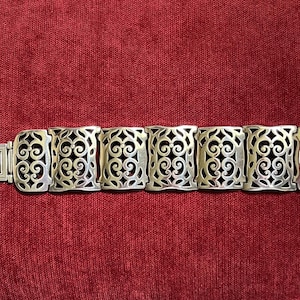 May include: A silver-toned link bracelet featuring seven rectangular segments. Each segment displays an intricate, openwork scroll pattern. The bracelet includes a rectangular clasp mechanism on one end and a flat metal tab on the other end.
