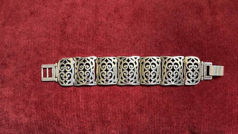 May include: A silver-toned link bracelet featuring seven rectangular segments. Each segment displays an intricate, openwork scroll pattern. The bracelet includes a rectangular clasp mechanism on one end and a flat metal tab on the other end.