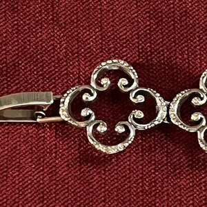 May include: A silver-toned metal bracelet featuring a series of repeating openwork quatrefoil links with a textured surface. The bracelet includes a polished metal box clasp mechanism on the end, connecting to the decorative filigree-style chain.