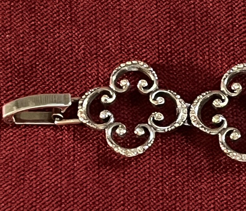 May include: A silver-toned metal bracelet featuring a series of repeating openwork quatrefoil links with a textured surface. The bracelet includes a polished metal box clasp mechanism on the end, connecting to the decorative filigree-style chain.