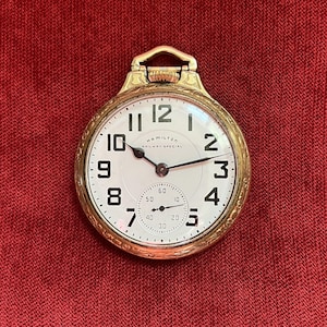 May include: A gold-toned Hamilton Railway Special pocket watch with a white face. The watch features black numerals, a small seconds dial, and the words "HAMILTON RAILWAY SPECIAL". The watch is set against a red background.