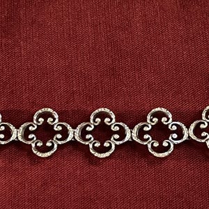 May include: A silver-toned metal bracelet featuring a repeating pattern of five interconnected quatrefoil-shaped links. Each link displays a detailed scrollwork design with a textured surface, creating a continuous chain of symmetrical floral-inspired motifs.