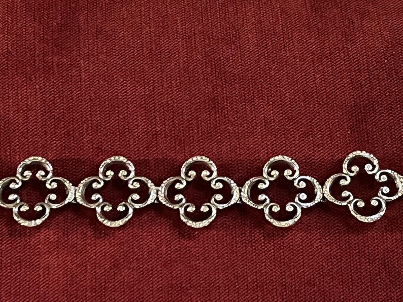 May include: A silver-toned metal bracelet featuring a repeating pattern of five interconnected quatrefoil-shaped links. Each link displays a detailed scrollwork design with a textured surface, creating a continuous chain of symmetrical floral-inspired motifs.