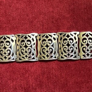 May include: A silver-toned metal link bracelet featuring a series of rectangular segments. Each segment displays an intricate, openwork filigree pattern of symmetrical scrolls and curves. The bracelet concludes with a flat, rectangular metal clasp.