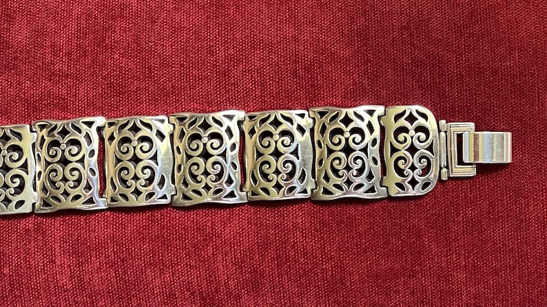 May include: A silver-toned metal link bracelet featuring a series of rectangular segments. Each segment displays an intricate, openwork filigree pattern of symmetrical scrolls and curves. The bracelet concludes with a flat, rectangular metal clasp.