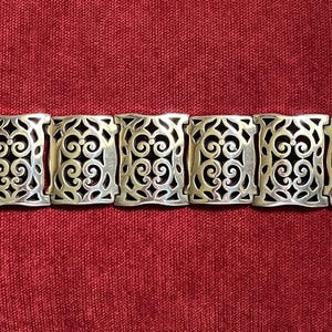 May include: A silver-toned metal bracelet featuring seven rectangular links with an intricate openwork scroll pattern. The bracelet includes a flat, rectangular clasp mechanism on each end, providing a secure closure for the linked design.