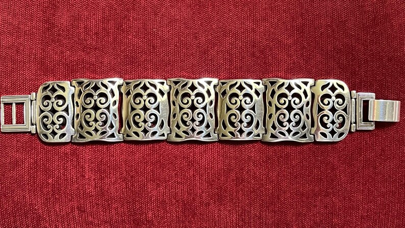 May include: A silver-toned metal bracelet featuring seven rectangular links with an intricate openwork scroll pattern. The bracelet includes a flat, rectangular clasp mechanism on each end, providing a secure closure for the linked design.