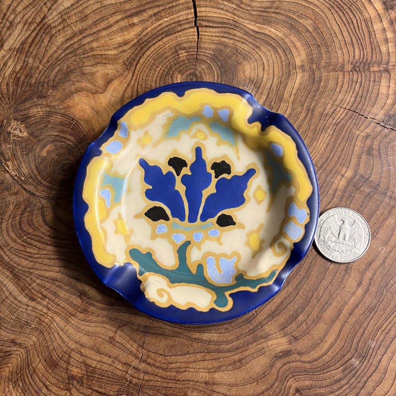 May include: A round, ceramic ashtray with a blue, yellow, and cream floral design. The ashtray has a dark blue rim and a central floral motif in blue and black. A US quarter is next to the ashtray for size comparison.