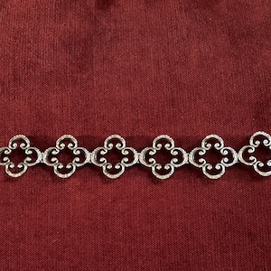 May include: A silver-toned metal bracelet featuring six repeating openwork quatrefoil links connected in a row. The design includes intricate scrollwork details within each clover-shaped link, finished with a simple rectangular clasp mechanism.
