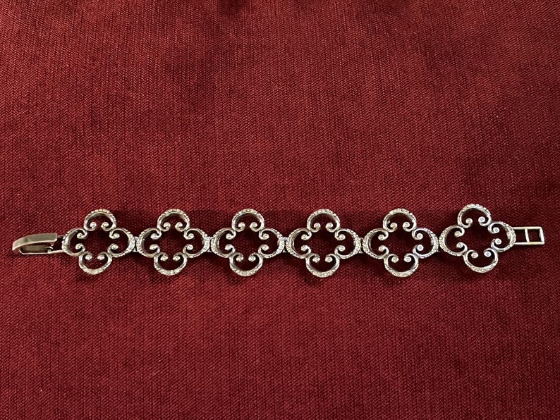 May include: A silver-toned metal bracelet featuring six repeating openwork quatrefoil links connected in a row. The design includes intricate scrollwork details within each clover-shaped link, finished with a simple rectangular clasp mechanism.