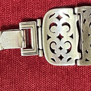 May include: A silver-toned metal bracelet featuring a series of rectangular links with an intricate cut-out filigree pattern. The bracelet includes a secure hinged clasp mechanism with the brand name Brighton engraved on the metal surface.