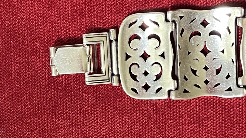 May include: A silver-toned metal bracelet featuring a series of rectangular links with an intricate cut-out filigree pattern. The bracelet includes a secure hinged clasp mechanism with the brand name Brighton engraved on the metal surface.