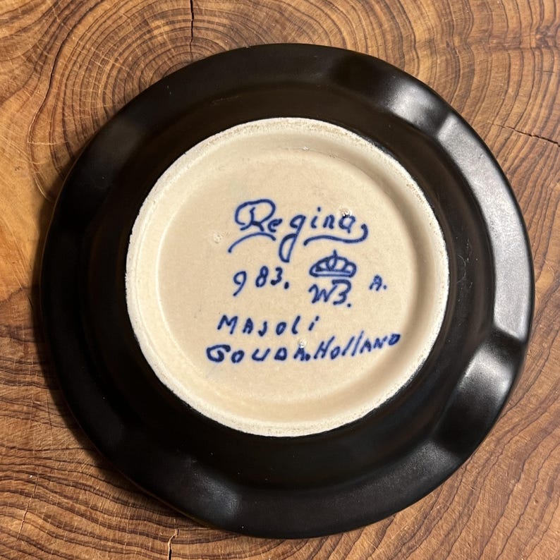 May include: A black ceramic ashtray with a light beige interior. The interior features blue text that reads "Regina", "983, W3, A", and "Majoli Gouda Holland". The ashtray is circular and sits on a wooden surface.