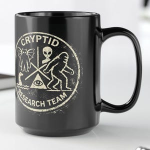 May include: Black ceramic mug with a white Cryptid Research Team graphic. The design features a bat-like creature, an alien, a Bigfoot, an eye, and a boat. The mug is on a white surface.