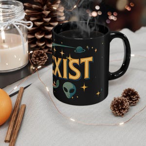 May include: Black ceramic mug with the word "EXIST" in yellow and blue, along with alien and space-themed graphics. The mug has a curved handle and is filled with a dark liquid, possibly coffee or tea.