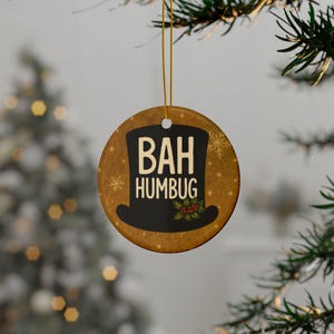 May include: A round Christmas ornament with a gold string for hanging. The ornament has a brown background with snowflakes and the words "BAH HUMBUG" in white. A black top hat graphic is in the center, with holly and berries.