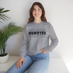 May include: A heather gray sweatshirt with the word "MOMSTER" in a layered, dripping font. The word is in black and gray. The sweatshirt has long sleeves and a crew neck.