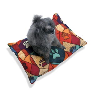 Puede incluir: Un caniche gris descansa sobre una colorida cama para perros. La cama presenta un diseño de patchwork con huellas de patas en tonos azules, naranjas, rojos y morados. La cama rectangular parece suave y cómoda.