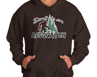 May include: A dark brown hooded sweatshirt with a graphic design. The design features a silhouette of Bigfoot, pine trees, and the text "Don't be an ASSQUATCH" in white and green. The sweatshirt has a front pocket.