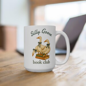 May include: White ceramic mug featuring the text "Silly Goose book club." The mug has a whimsical illustration of three geese wearing green bows, standing on books. The mug has a large handle.