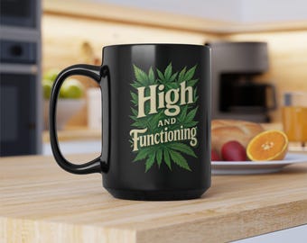 May include: Black ceramic mug with the words "High and Functioning" in a green and cream design. The text is surrounded by green cannabis leaves. The mug has a large handle and is sitting on a wooden surface.