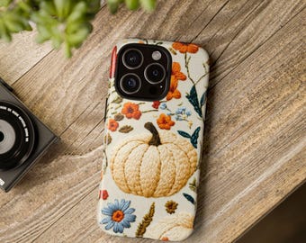 May include: A beige phone case with an embroidered pumpkin and floral design. The case features a large pumpkin in the center, surrounded by orange, blue, and green flowers and leaves. The case is on a wooden surface.