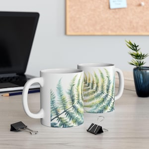 May include: Two white ceramic mugs with a watercolor fern design. The mugs are on a wooden table with a laptop, pencils, and a plant in the background.