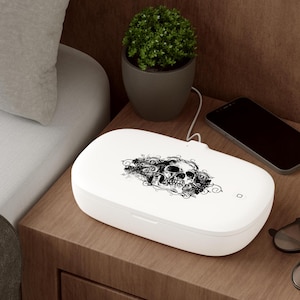 May include: White UV sanitizer with a skull and floral design on the lid. The sanitizer is plugged in and sitting on a wooden bedside table next to a smartphone and glasses.