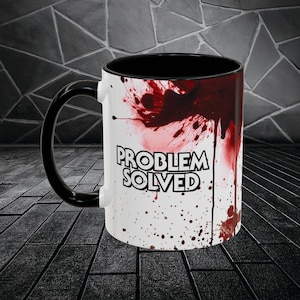 Peut inclure: Mug en céramique blanche avec une anse et un intérieur noirs. Le mug présente un motif d'éclaboussures rouges et le texte "PROBLEM SOLVED" en lettres blanches en gras. Le mug est posé sur un fond sombre.