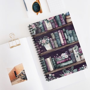 May include: A spiral-bound notebook featuring a watercolor illustration of a bookshelf filled with books and floral arrangements. The books are in shades of purple, green, and cream. The notebook is open on a white surface.
