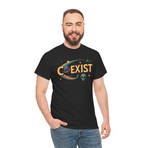 May include: Black t-shirt featuring the word "COEXIST" in a colorful, stylized font with a space-themed graphic. The design includes planets, stars, and an alien head. The shirt is made of a soft material.