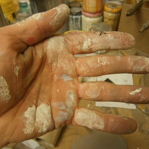May include: A close-up of a hand covered in white paint. The hand is open and the fingers are spread out. The paint is thick and uneven, and there are several spots where the paint has dried and cracked.