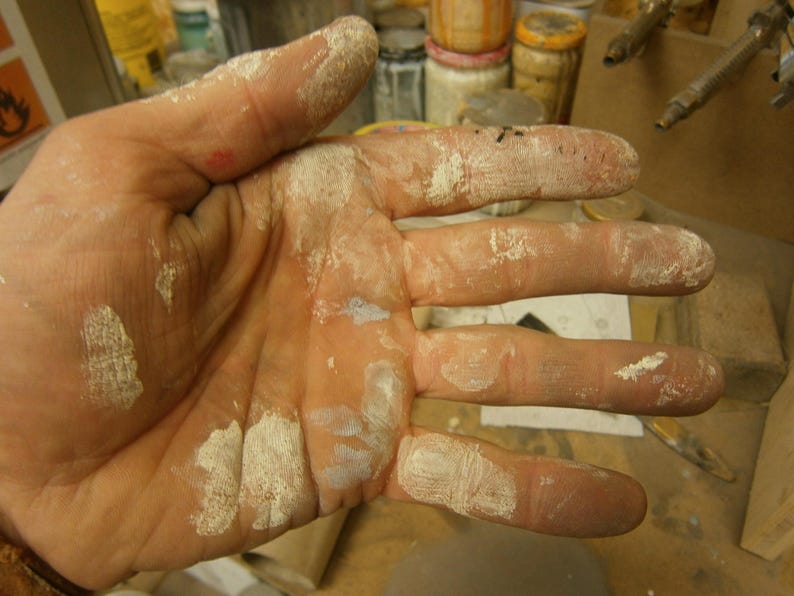 May include: A close-up of a hand covered in white paint. The hand is open and the fingers are spread out. The paint is thick and uneven, and there are several spots where the paint has dried and cracked.