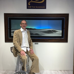 May include: A man in a tan suit sits on a stool in front of a framed sand art piece. The artwork is lit from behind with blue light. The frame is dark wood with a black and gold inlay. The artwork is titled "KB The Sandman".