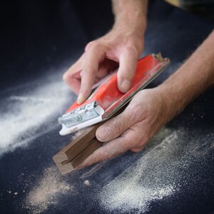 May include: A person's hands sanding a piece of wood with a red and white power sander. The wood is dark brown and has a groove in the center. There is sawdust on the surface.