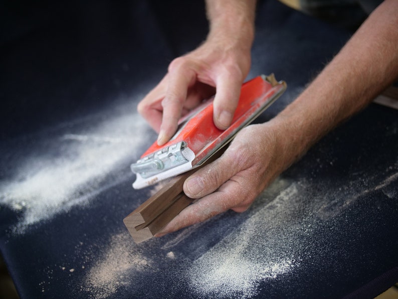 May include: A person's hands sanding a piece of wood with a red and white power sander. The wood is dark brown and has a groove in the center. There is sawdust on the surface.