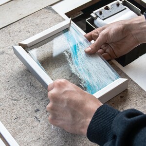 May include: A person is holding a white frame with a glass panel inside. The glass panel has a blue and white abstract design.