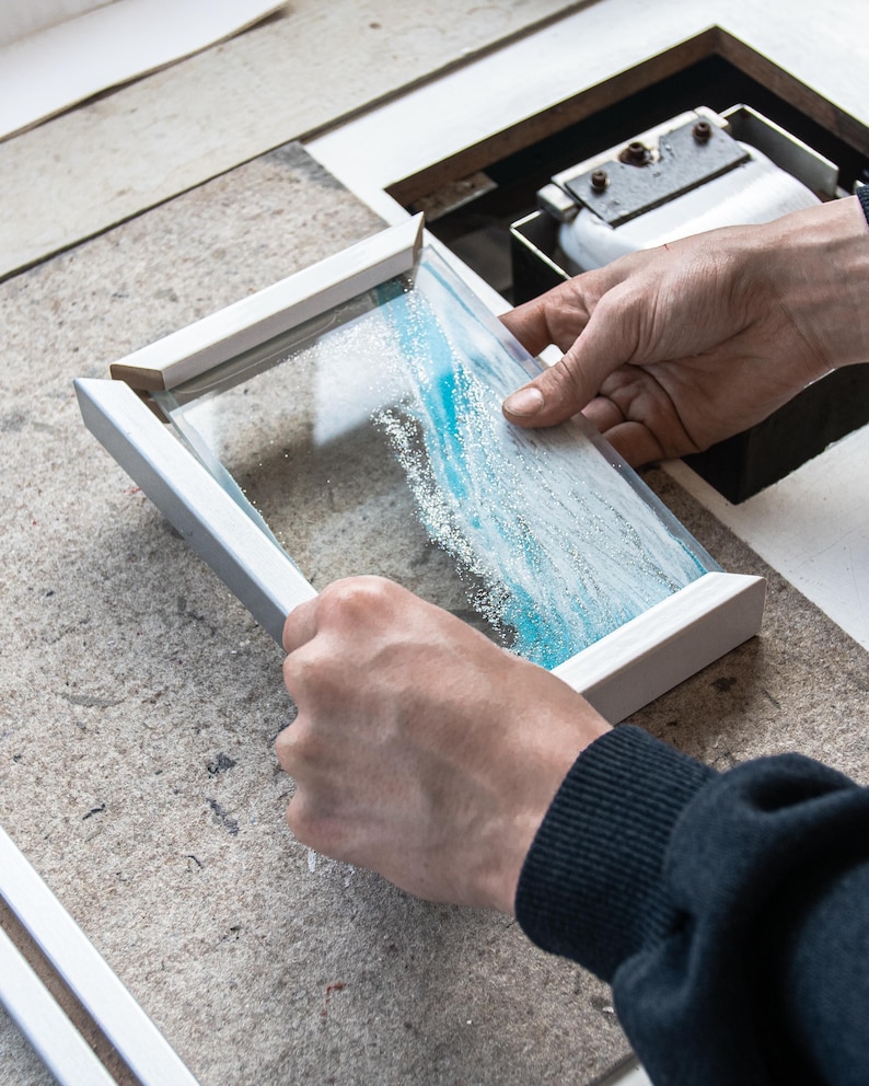 May include: A person is holding a white frame with a glass panel inside. The glass panel has a blue and white abstract design.